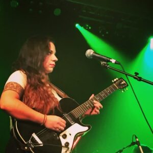 About Meghan is playing a black and white guitar on stage bathed in green spotlights with her long black hair cascading over her shoulders